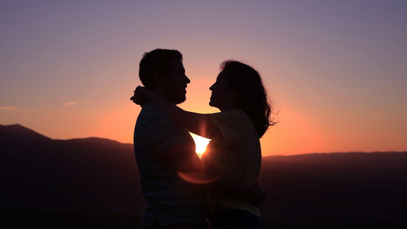 Silhouet van een man en een vrouw die elkaar omarmen en aankijken, met de ondergaande zon tussen hen in tegen een paarse avondlucht